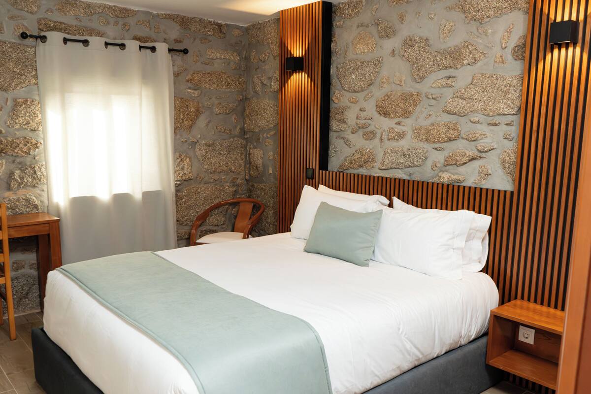 Un lit avec une tête de lit, une table de chevet en bois, une chaise et un mur en pierre.