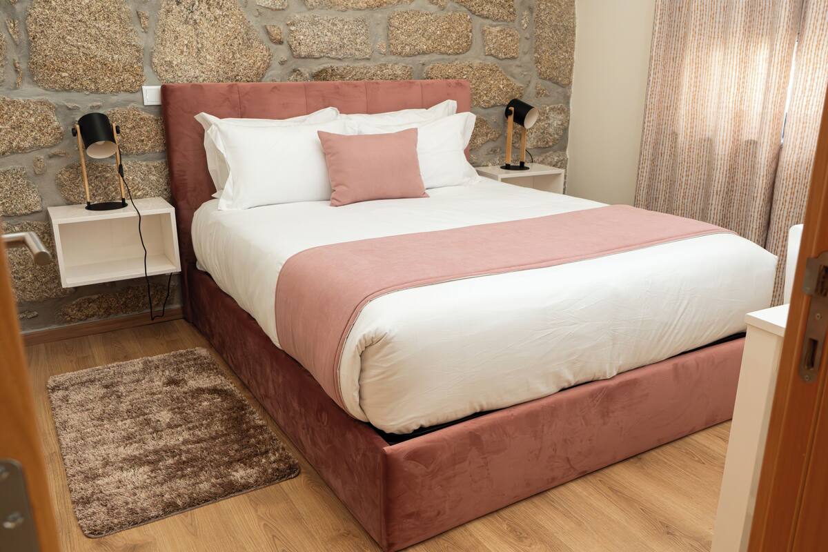 Une chambre avec un mur en pierre, un lit avec une literie blanche et rose, deux tables de chevet avec des lampes et un plancher en bois.