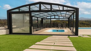 Una estructura moderna con paredes de vidrio, una piscina, un patio con sillas y un camino de piedra que conduce a ella.