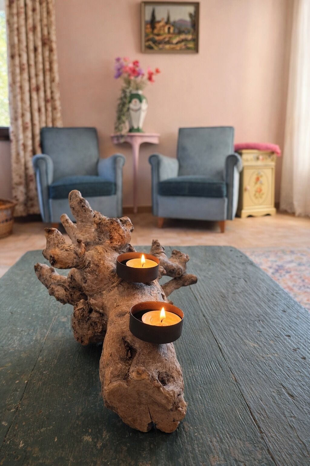 Un tavolo rustico in legno con tre candele, due poltrone blu, un tavolino con un vaso di fiori e un quadro appeso al muro.