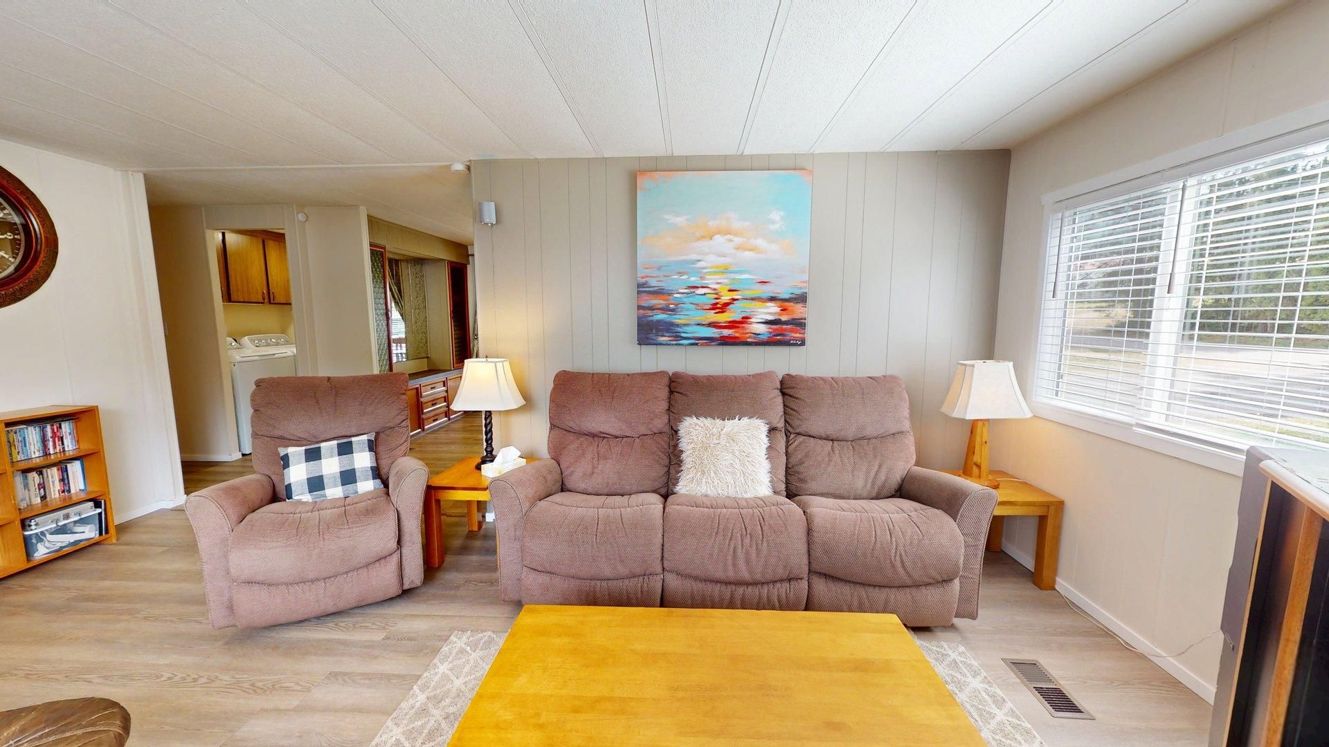 A living room with a sofa, armchairs, a coffee table, a painting on the wall, a bookshelf, and a window with blinds.