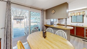 Quarto com mesa de jantar de madeira, cadeiras brancas, área de cozinha com micro-ondas e vista através de uma porta de vidro.