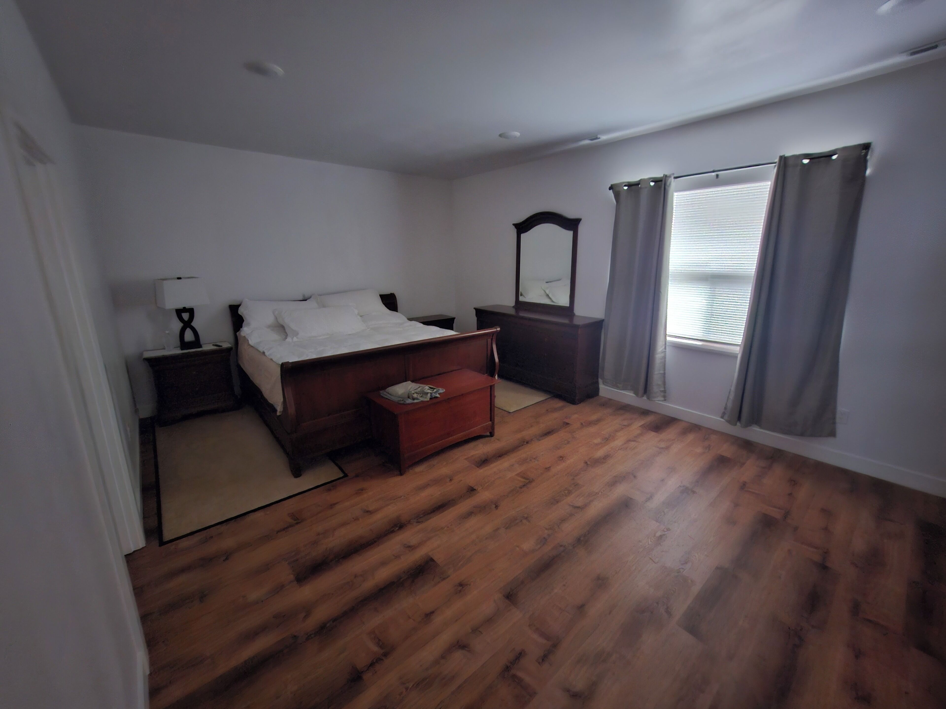 Ein Schlafzimmer mit Holzboden, einem Bett mit weißer Bettwäsche, einem Nachttisch, einer Kommode, einem Spiegel und einem Fenster mit Vorhängen.