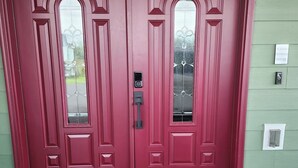 A red double door with a glass panel, a doormat, and a 'Please Remove Shoes' sign on the wall.