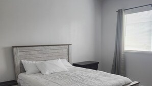A bedroom with a wooden bed, white bedding, a nightstand, and a window with curtains.