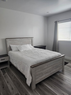 A bedroom with a wooden bed, white bedding, a nightstand, and a window with curtains.