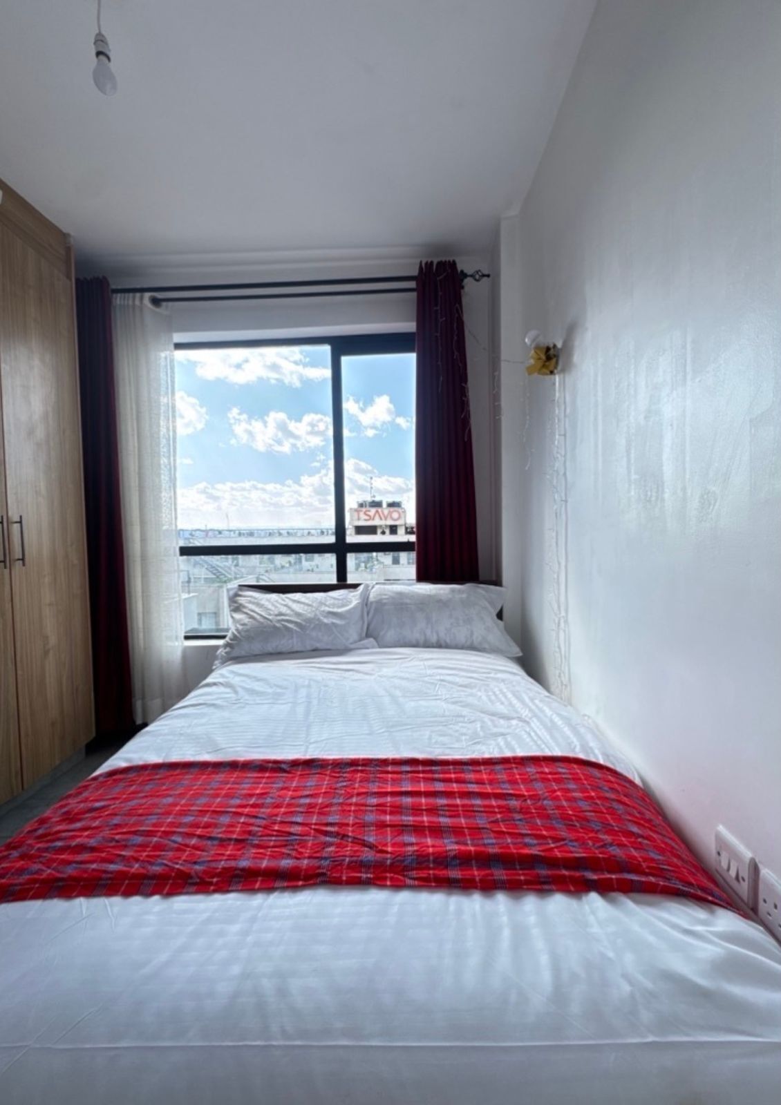 Een slaapkamer met een bed, een raam met gordijnen, uitzicht op de lucht en een stadsgezicht.