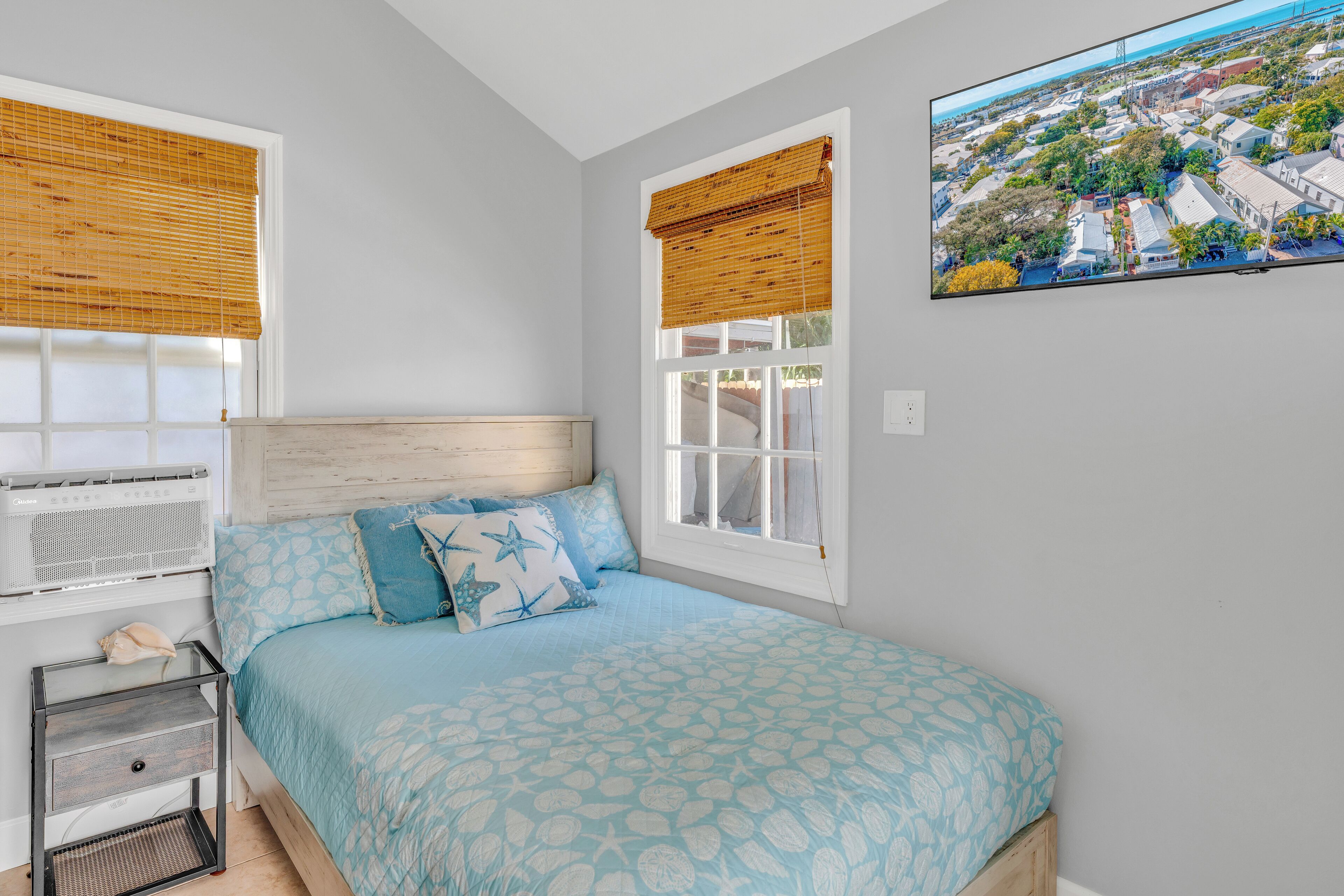 A bedroom with a bed, two windows with blinds, a nightstand, and a wall-mounted TV.