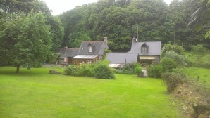 Une maison de campagne avec un toit en pente, entourée d’un champ herbeux et d’une forêt dense.