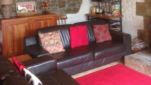 Un salon chaleureux avec un mur en pierre, un plafond en bois, un canapé en cuir agrémenté de coussins rouges et une petite table sur laquelle sont disposés divers objets.