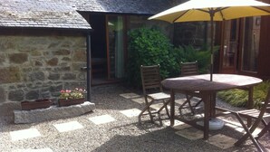 Un bâtiment en pierre avec une terrasse en gravier, une table en bois entourée de chaises et un parasol jaune.