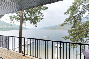 Um deck de madeira com guarda-corpo preto, com vista para um lago calmo, cercado por árvores e montanhas ao longe.