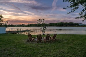 A lakeside view with chairs facing the water, a grassy area, and a building in the background. - Lakefront Family Escape with Pool | Near Disney (Davenport)