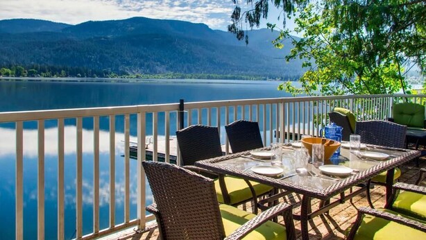 A dining area with a table set for two, overlooking a lake with mountains in the background. - Aussie Summer (Christina Lake)