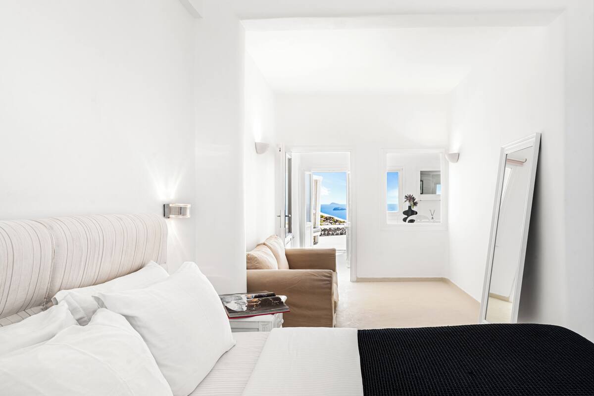 A hotel room with a bed, a bedside table with books, a sofa, a mirror, and a view of the outdoors.