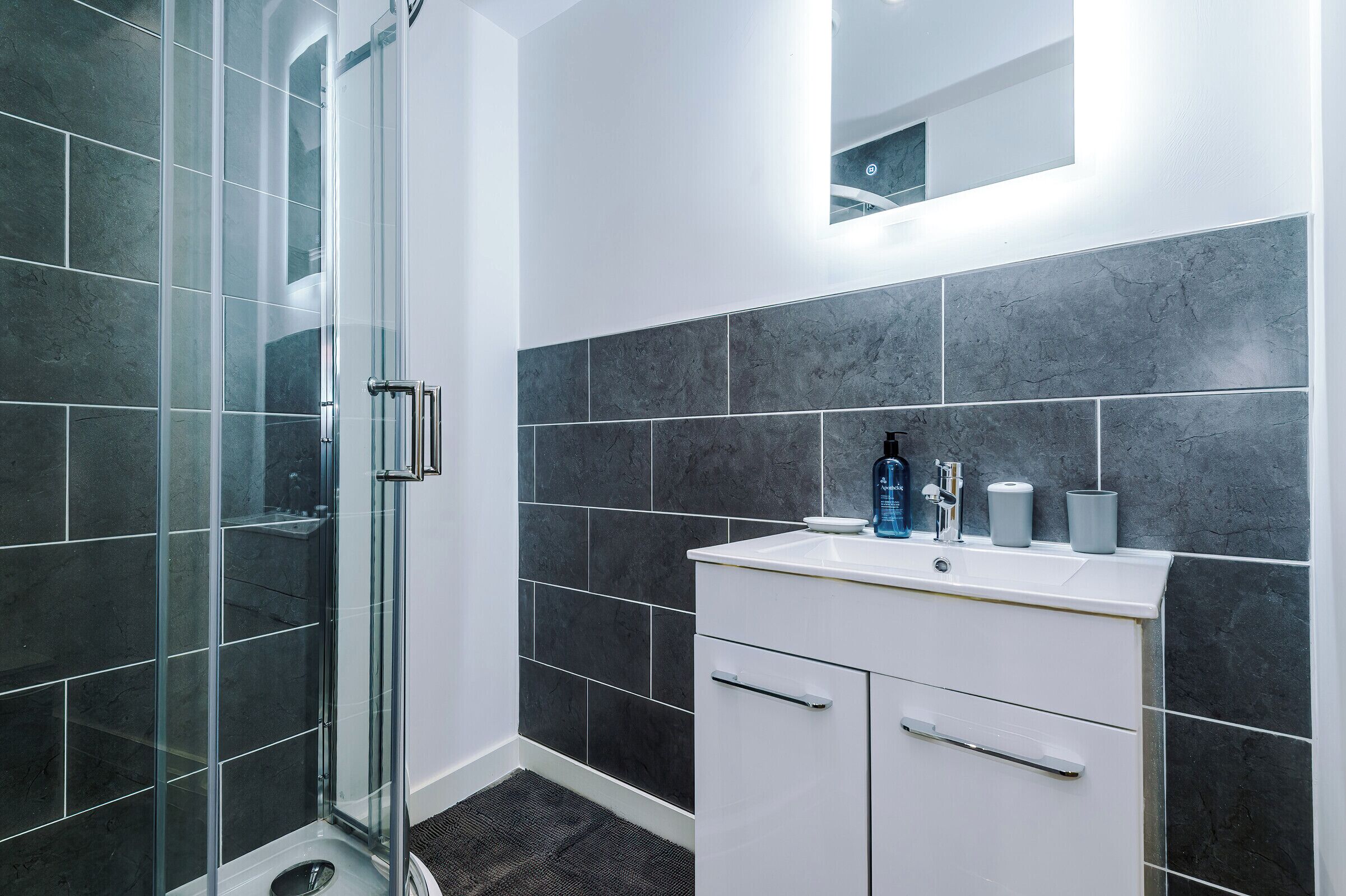 A modern bathroom with a glass shower enclosure, dark tiled walls, a white vanity with a mirror, and a small sink.