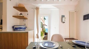 A dining area with a table set for two, a blue and white plate, a bowl, and a spoon. There are wooden shelves above the counter and a view of a building outside. - Beach 1Min | Sea View| 2BR| A/C| by Tilo (Genoa)