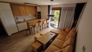 A modern kitchen with wooden cabinets, a dining area with a sofa and chairs, and a view of the outdoors through large windows. - Apartment, 2BDR, shared pool (Sarajevo)