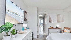 Ett hotellrum med en säng, ett skrivbord med en lampa, en tv som visar en strandscen och en hall som leder till ett annat rum.