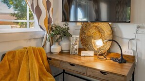 A room with a wooden desk, a television mounted on the wall, a patterned lampshade, and a window with curtains. - Boho Blue Heron, BBH 7532 (Birch Bay)