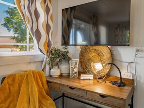 A room with a wooden desk, a television mounted on the wall, a patterned lampshade, and a window with curtains.
