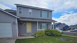 A two-story residential building with a front garage, a small front yard with grass and a bush, and a clear sky. - Spacious Family Home w Garage  Gladys Retreat (Hermiston)