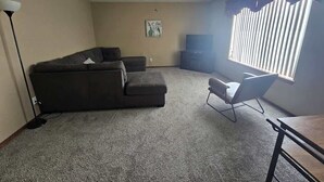 A living room with a dark-colored sofa, a single chair, a ceiling fan, and a window with blinds. - Spacious Family Home w Garage  Gladys Retreat (Hermiston)