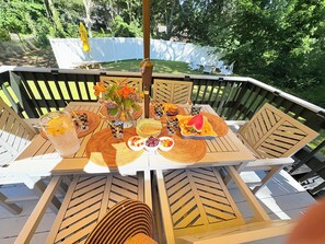 Ein runder Tisch im Freien mit weißem Tischtuch, umgeben von Holzstühlen mit Querstreben, auf einer Terrasse mit weißem Zaun im Hintergrund.