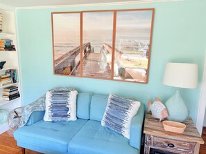 Ein Zimmer mit einem hellblauen Sofa, einem Holznachttisch mit Lampe, einem Bücherregal und einem großen gerahmten Bild mit einer Strandansicht.