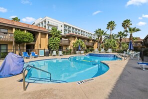 Zona de piscina con sillas de descanso y sombrillas, un cartel que indica 'no saltos, 3 pies' y un edificio con balcones y palmeras.