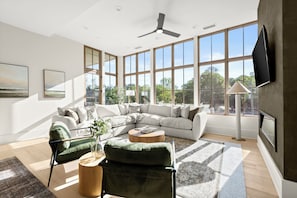 Un salon moderne avec un canapé d’angle, un fauteuil vert, une table basse ronde et une grande fenêtre donnant sur la verdure.