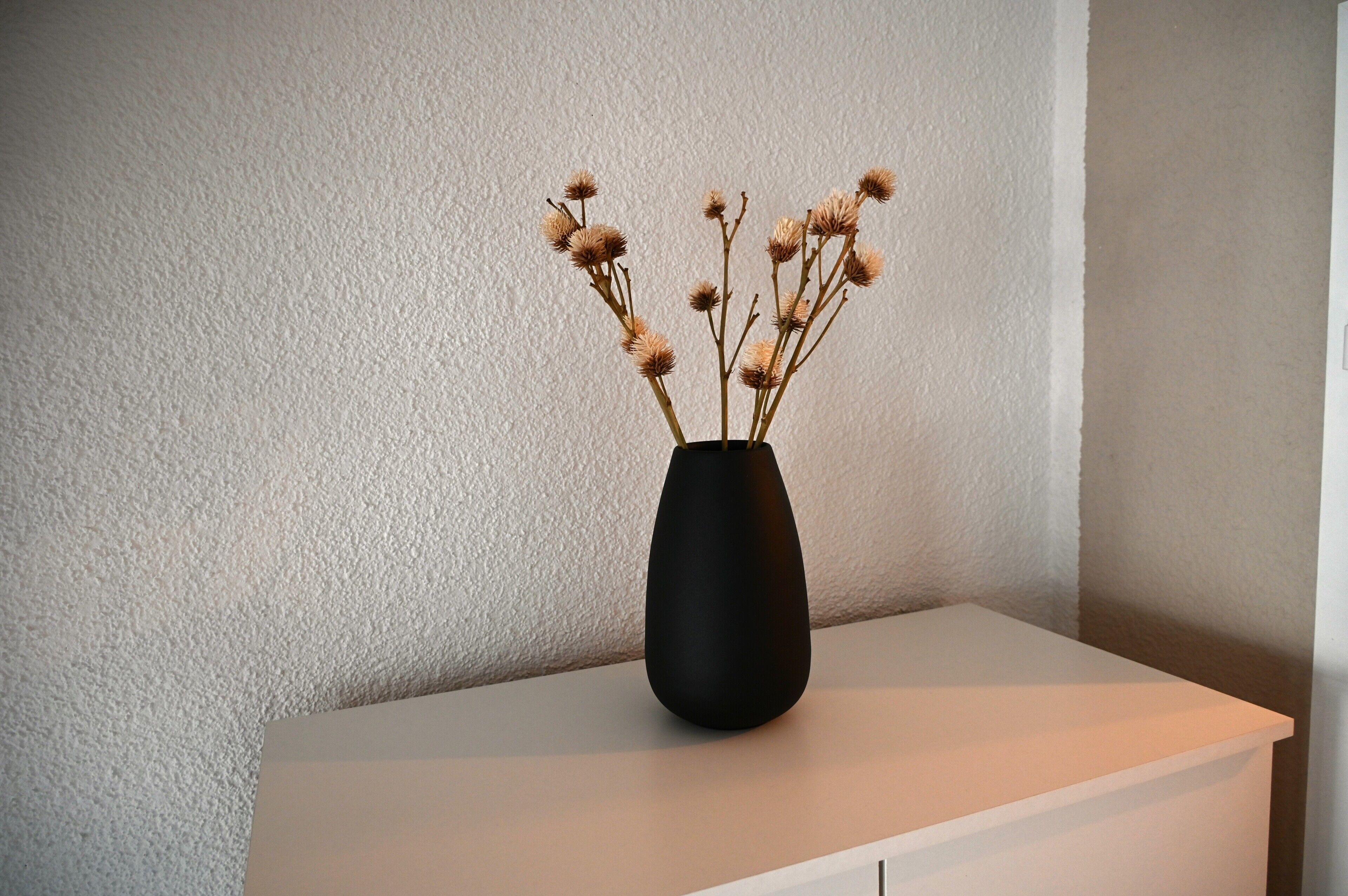 Un vase noir contenant des fleurs séchées, posé sur une table blanche, le tout sur fond d’un mur à la texture marquée.
