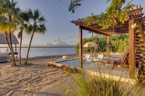 Un espace piscine aménagé au bord de la plage, avec des chaises longues, une pergola et une structure à toit de chaume.