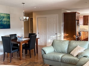 A dining area with a wooden table and chairs, a light-colored sofa, and a kitchen with wooden cabinets. - Garden level suite in sunny Osoyoos. Minutes from beaches, parks and golf! (Osoyoos)