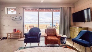 Ein Zimmer mit Deckenventilator, Fernseher, großem Fenster mit Blick auf den Strand und zwei Stühlen.