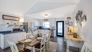 A spacious kitchen with white cabinetry, a dining area with a wooden table and chairs, and a view of the living area through an open door. - Oceanfront, pet friendly, fenced yard (Oak Island)