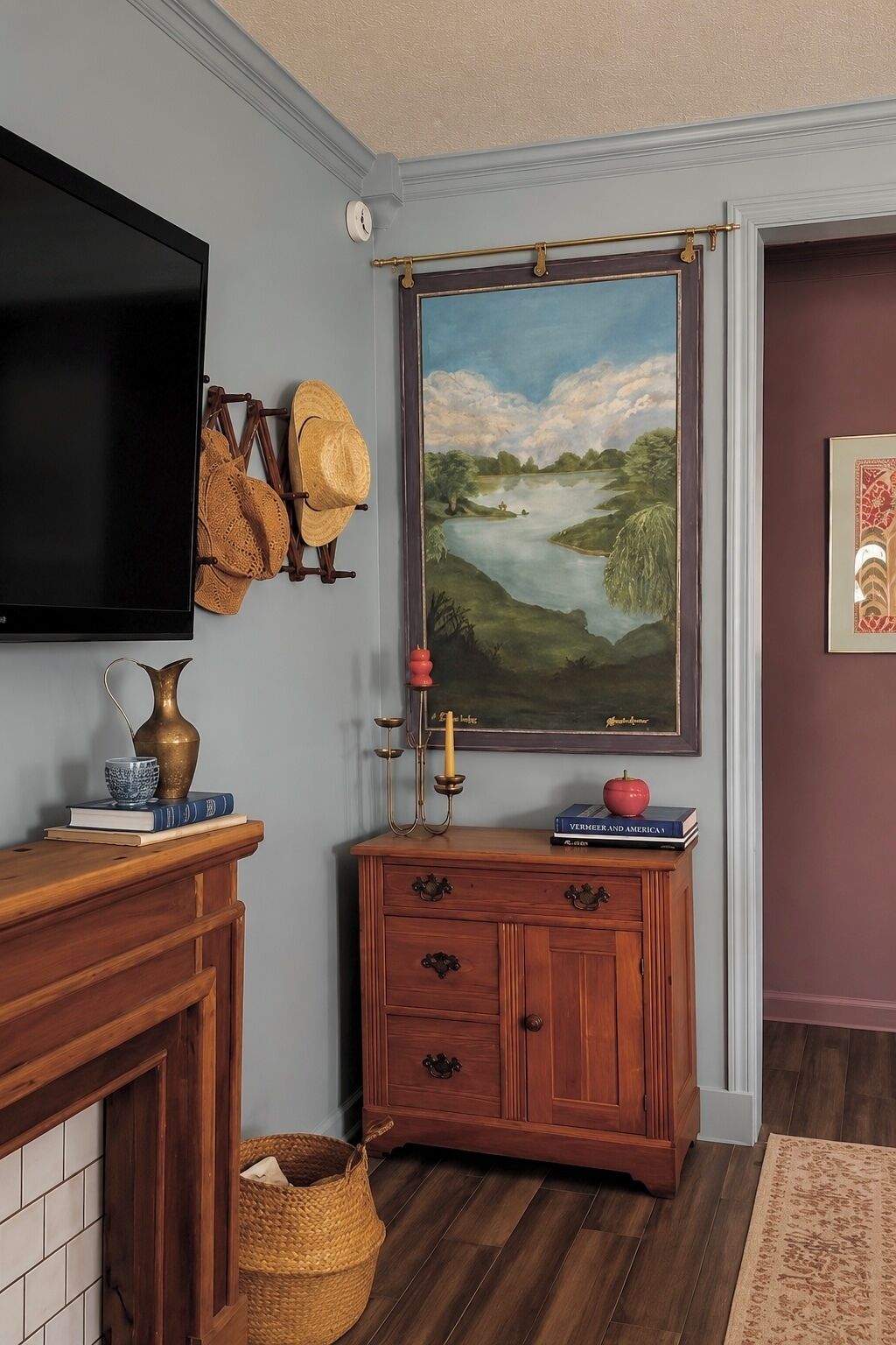 Een kamer met een wandgemonteerde flatscreen-tv, een houten kast met lades, een rieten mand en een schilderij van een landschap.
