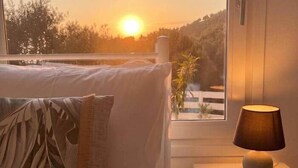 Un dormitorio con cama, mesita de noche con lámpara y vistas a la puesta de sol a través de una ventana.