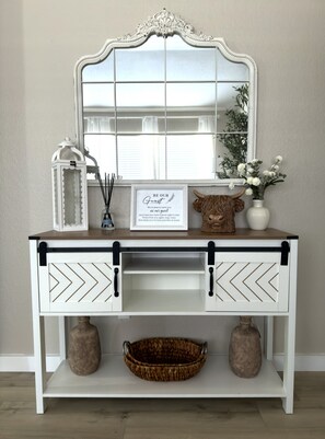 Une console blanche avec un miroir, un panneau décoratif encadré, des objets décoratifs et deux paniers.