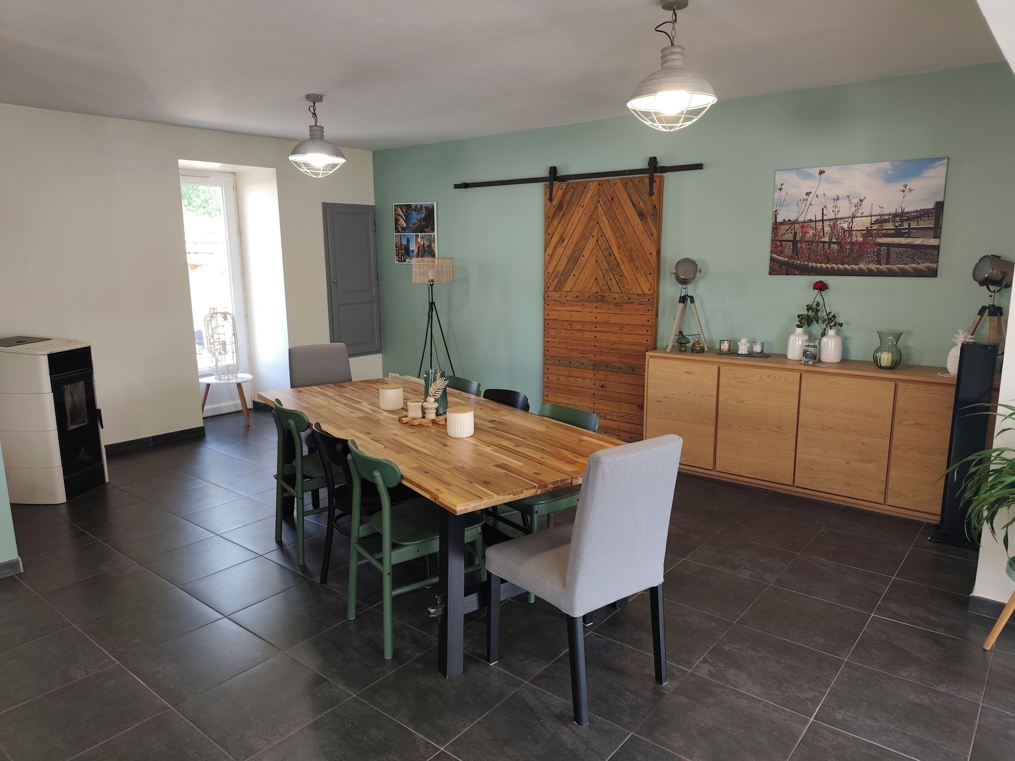 Un coin repas avec une table en bois, des chaises vertes, un meuble contenant des objets décoratifs et un tableau accroché au mur.