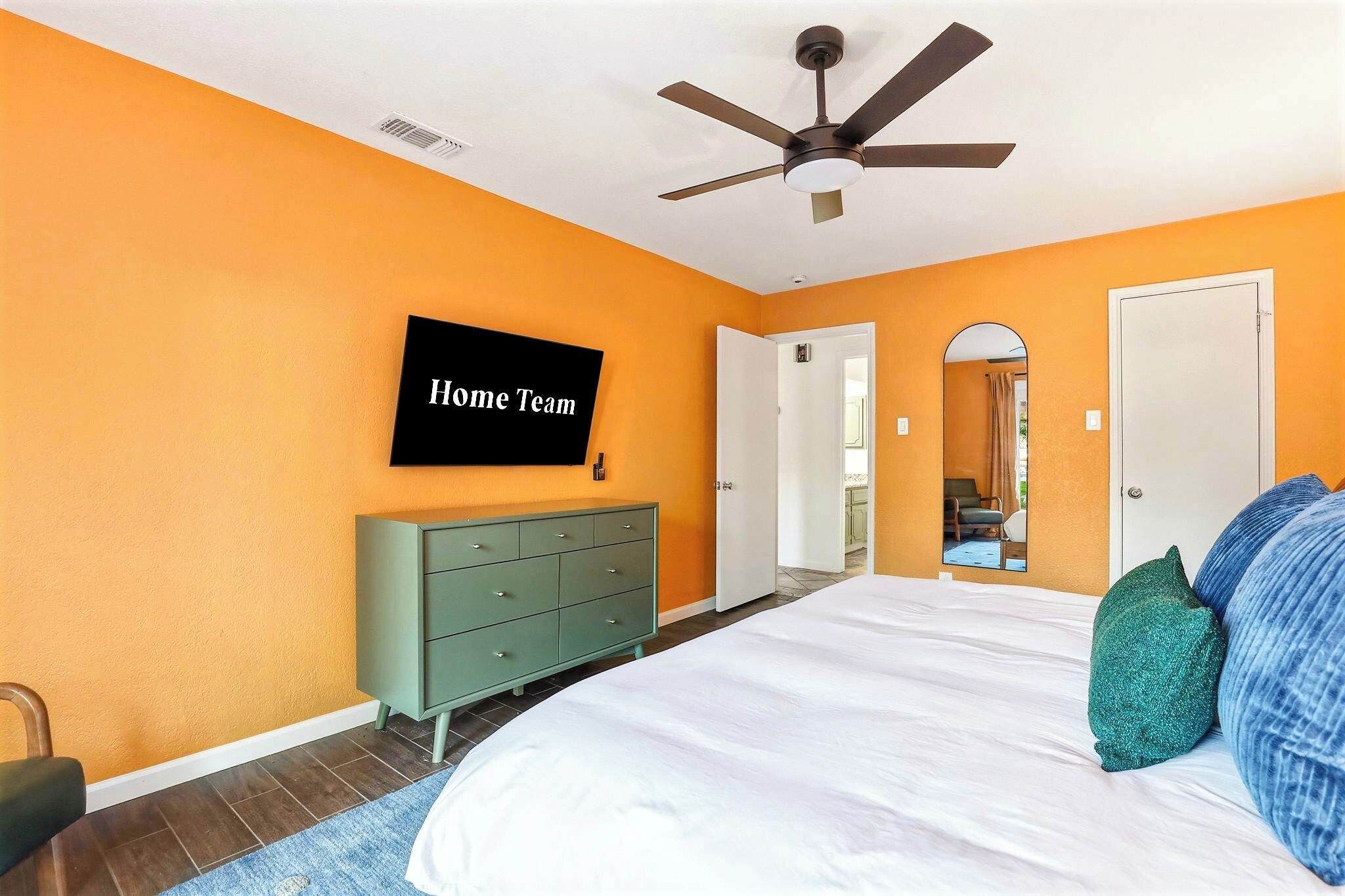 A bedroom with an orange wall, a bed with white bedding, a green dresser, a ceiling fan, and a TV displaying 'Home Team'.
