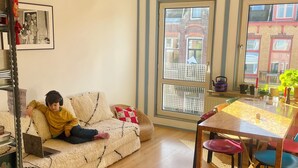 A cozy living room with a sofa, a dining table with chairs, a laptop on the sofa, and a large window with a view of buildings.