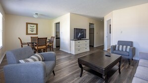 A living room with a sofa, armchair, coffee table, and a TV. There is a dining area with chairs and a table, and a kitchen area with cabinets and a refrigerator. - Keystone Garden Villa G (Indianapolis)