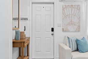 A white door with a handle, a wooden console table with baskets, a mirror, and a wall-mounted light switch. - Delray Beach Cottage • Jul/Aug Deal (Delray Beach)