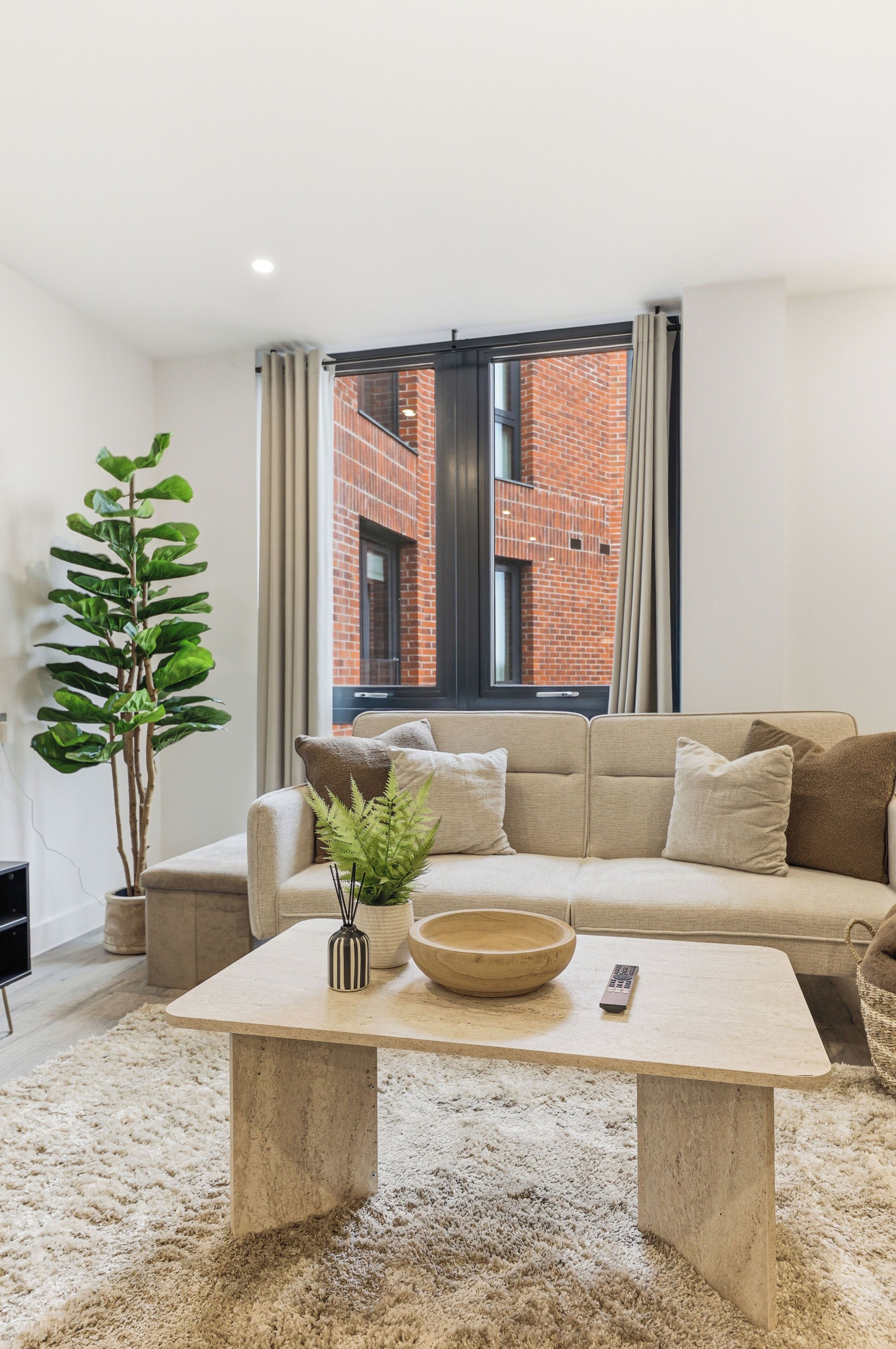 A modern living room with a beige sofa, a coffee table, a potted plant, and a view of a brick building through a window.