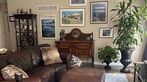 Ein Wohnzimmer mit braunem Ledersofa, einem Holztisch, einer großen Topfpflanze und gerahmten Bildern an der Wand.
