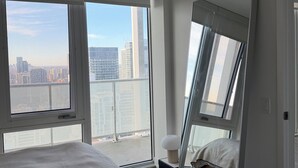 Un dormitorio con una cama grande, una mesita de noche, un espejo y vistas a un paisaje urbano a través de amplias ventanas.