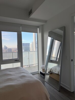 Ein Schlafzimmer mit einem großen Bett, einem Nachttisch, einem Spiegel und Blick auf eine Stadtkulisse durch große Fenster.