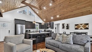 A spacious living area with a high ceiling, a kitchen with dark cabinets, and a gray sofa with patterned cushions. - Stone's Throw | Hot Tub + Fire Pit + Easy Roads (Blue Ridge)