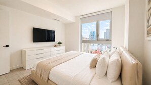 Ein Schlafzimmer mit einem Bett, einem an der Wand montierten Fernseher, einem Fenster mit Blick auf die Stadt und einer weißen Kommode.